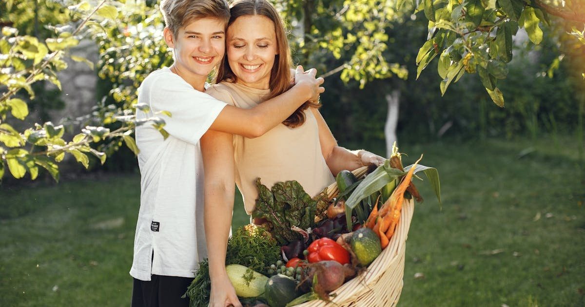 Mother and son smiling outdoors with a basket full of fresh vegetables, embracing in a sunny garden.
