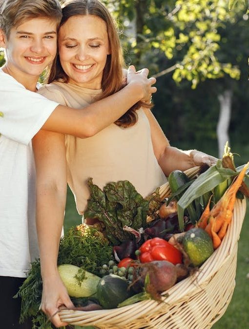 Mother and son smiling outdoors with a basket full of fresh vegetables, embracing in a sunny garden.