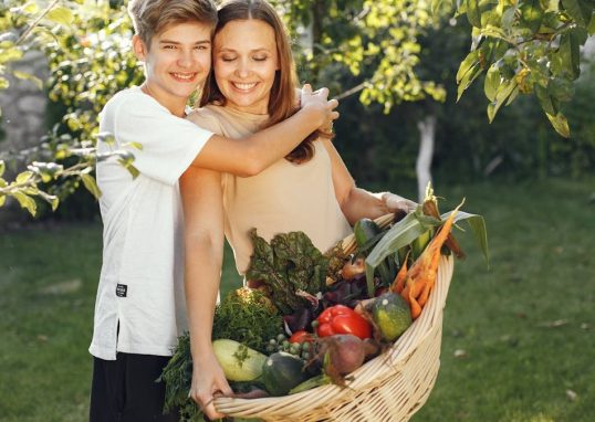 Mother and son smiling outdoors with a basket full of fresh vegetables, embracing in a sunny garden.