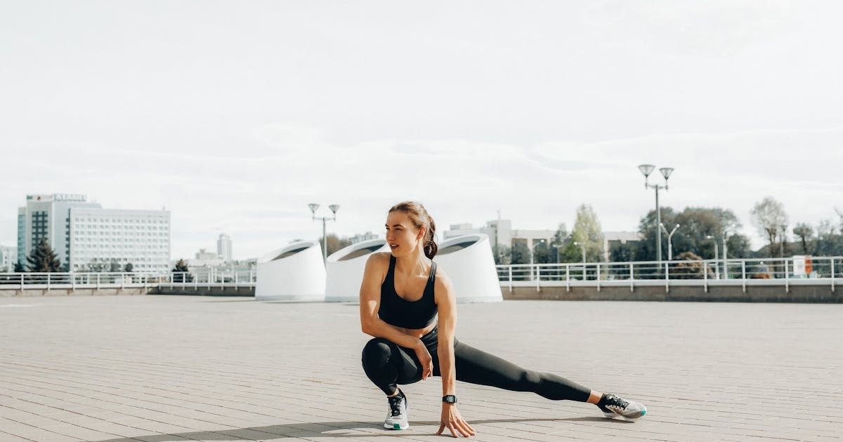Young woman in black sportswear stretching outdoors in a sunny urban environment.